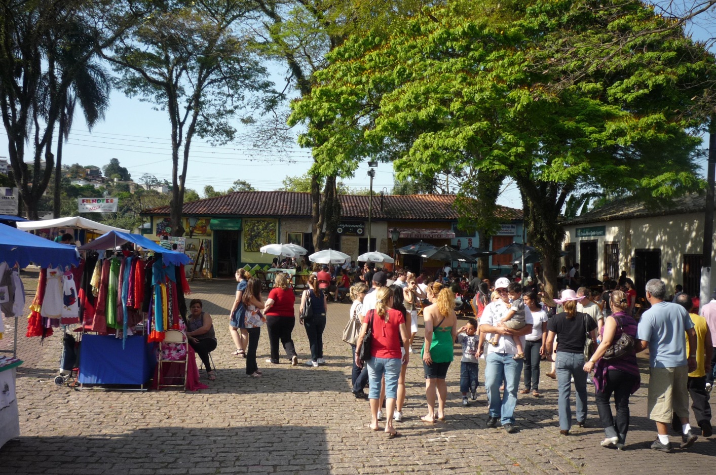 Barraca de roupas coloridas e visitantes caminhando por um largo arborizado durante a Feira de Artes de Embu das Artes.