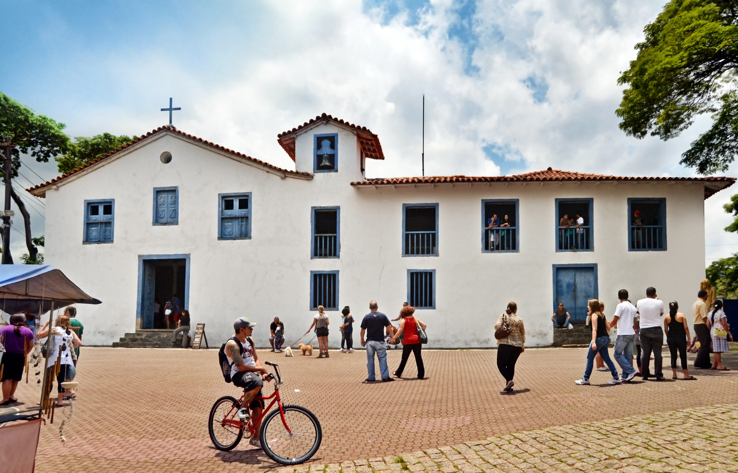 Fachada branca da Igreja de Nossa Senhora do Rosário, com janelas azuis e torre do sino, e pessoas no largo de pedra em frente.