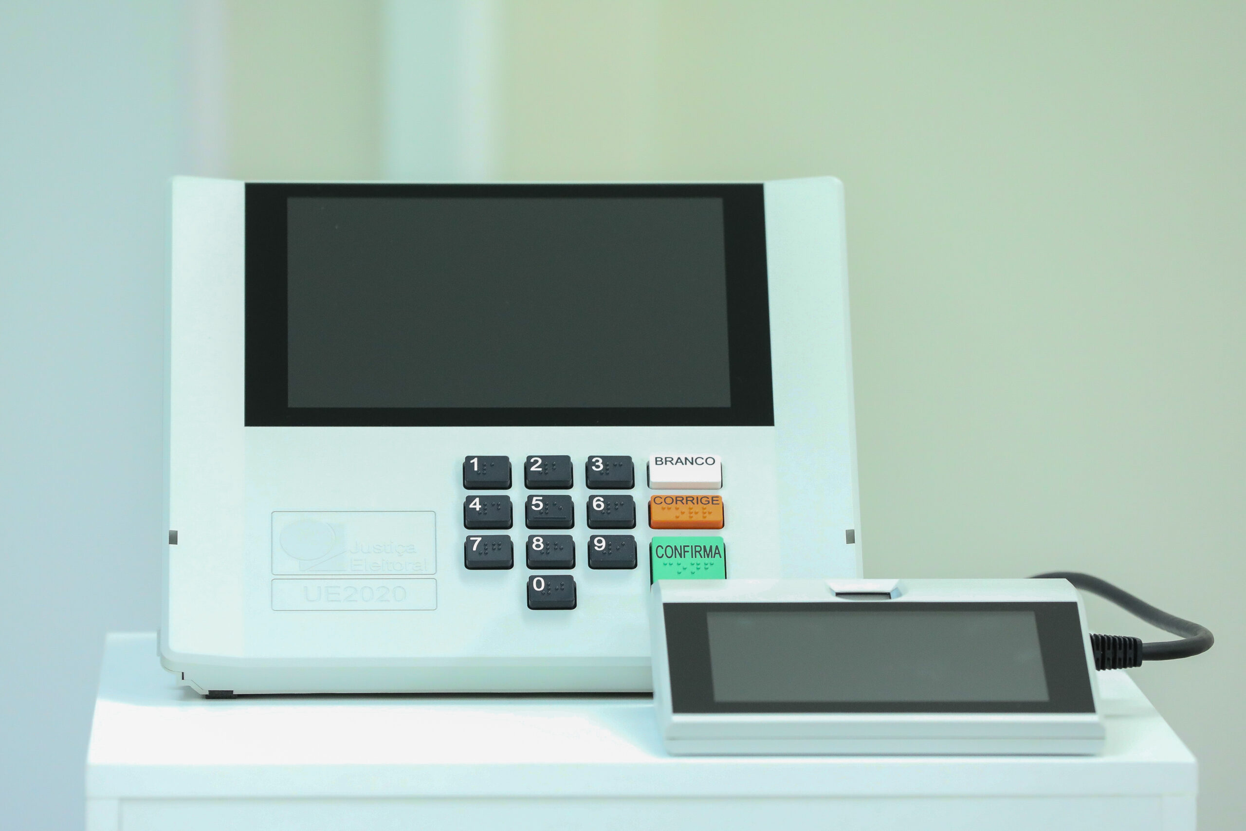 Urna eletrônica branca com teclado numérico e teclas Branco, Corrige e Confirma, ao lado do terminal do mesário, sobre uma mesa branca.